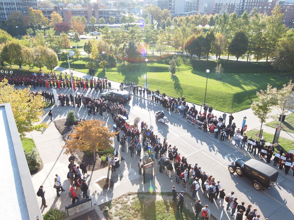 photo from on top of a building of a funeral procession with hundreds of people lining the street with two black trucks and a motorcycle escort