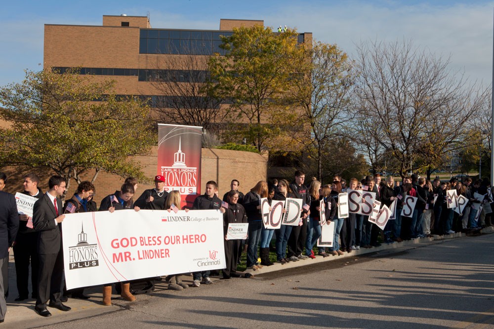 people line a road with a long white banner that says "God Bless our Hero Mr. Lindner" in front of a red brick building
