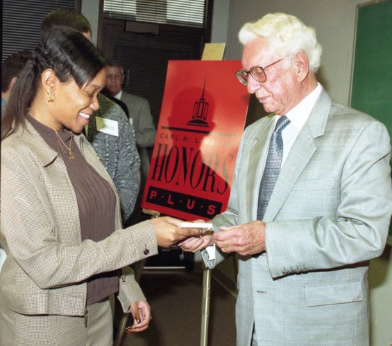 young woman wearing a brown blouse and light brown printed suit receives a deck of cards from man with white hair glasses white shirt, dark tie and dark grey suit in a classroom setting with the presence of other people in the background and a red decorative posterboard