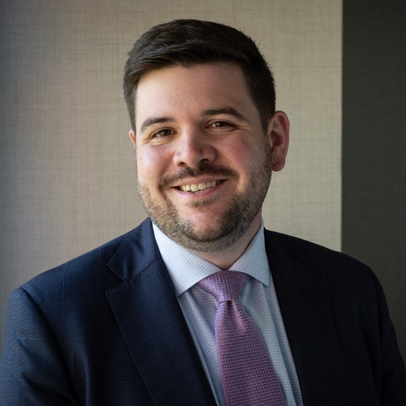 Man with dark short hair and a beard smiles for a headshot wearing a white shirt, purple tie and navy blue suit jacket