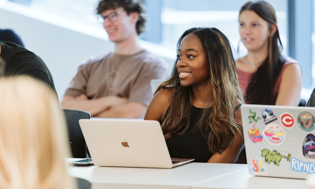 A student in a black shirt sits and smiles with her laptop in front of her.