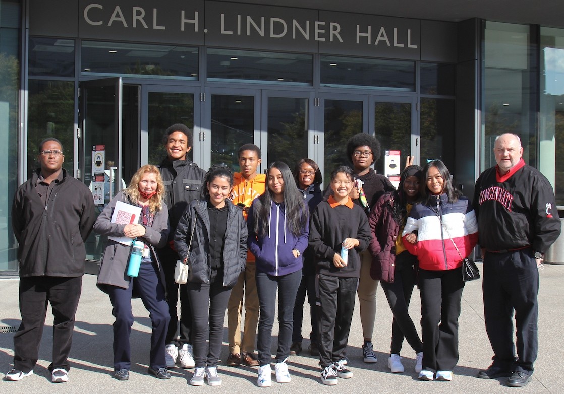 A group of about a dozen people, made up of high school students and teachers, stand outside Lindner Hall wearing jackets and coats on a sunny day