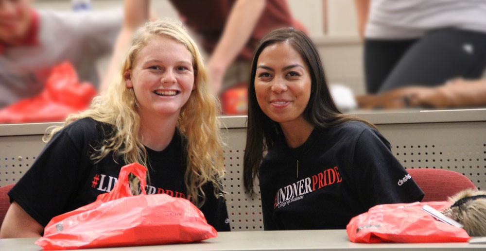 A girl with long blonde hair wearing a black shirt sits and smiles next to a girl with long dark hair weraing a black shirt at a long table with red bags on it