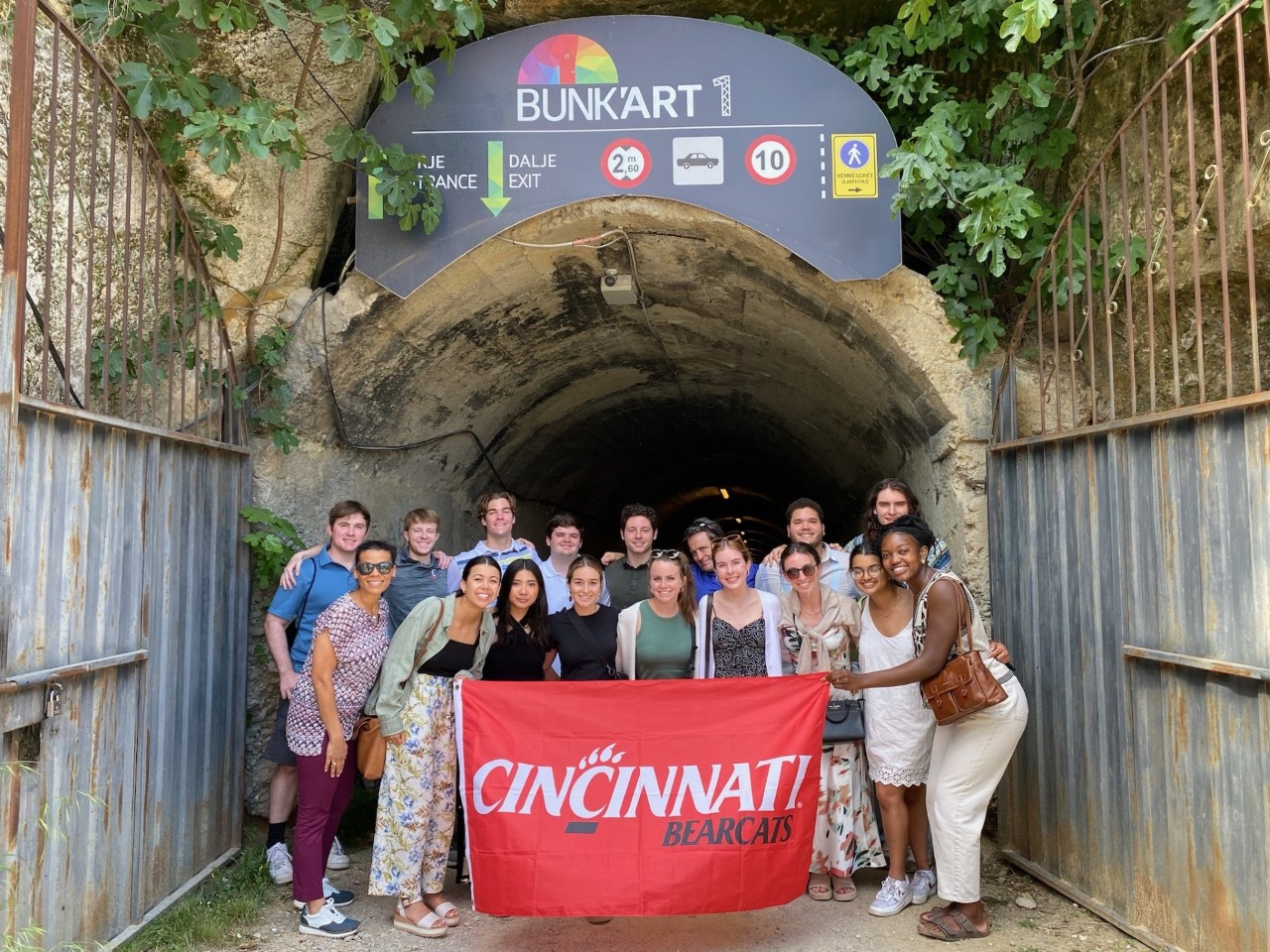 A group of 17 Lindner Business Honors students pose with a red Cincinnati Bearcats flag in front of a tunnel opening