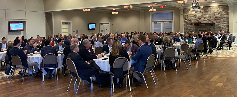 Attendees at a Real Estate Center Roundtable event sit at tables listening to a speaker standing on a stage at a podium with two screens to either side of them